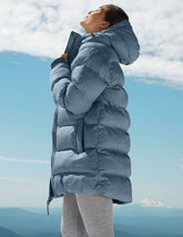woman wearing a blue puffer coat standing against a mountainous landscape with a clear sky.