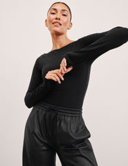 woman wearing a black long-sleeve boatneck bodysuit and black pants on a plain background.
