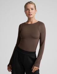 woman wearing a brown long-sleeve boat neck bodysuit and black pants against a plain background.