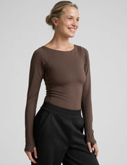 woman wearing a brown long-sleeve boat neck bodysuit and black pants against a plain background.