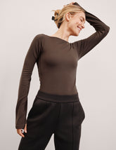woman wearing a brown long-sleeve boat neck bodysuit and black pants against a plain background.