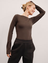 woman wearing a brown long-sleeve boat neck bodysuit and black pants against a plain background.