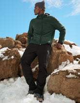 man wearing a green jacket, gray beanie, and black pants standing on snow-covered rocks with a blue sky.