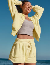 model in yellow jacket and shorts with gray top against a blue sky and ocean background.
