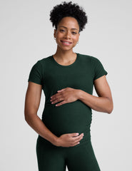 model woman wearing a dark green maternity t-shirt and leggings against a light gray background.