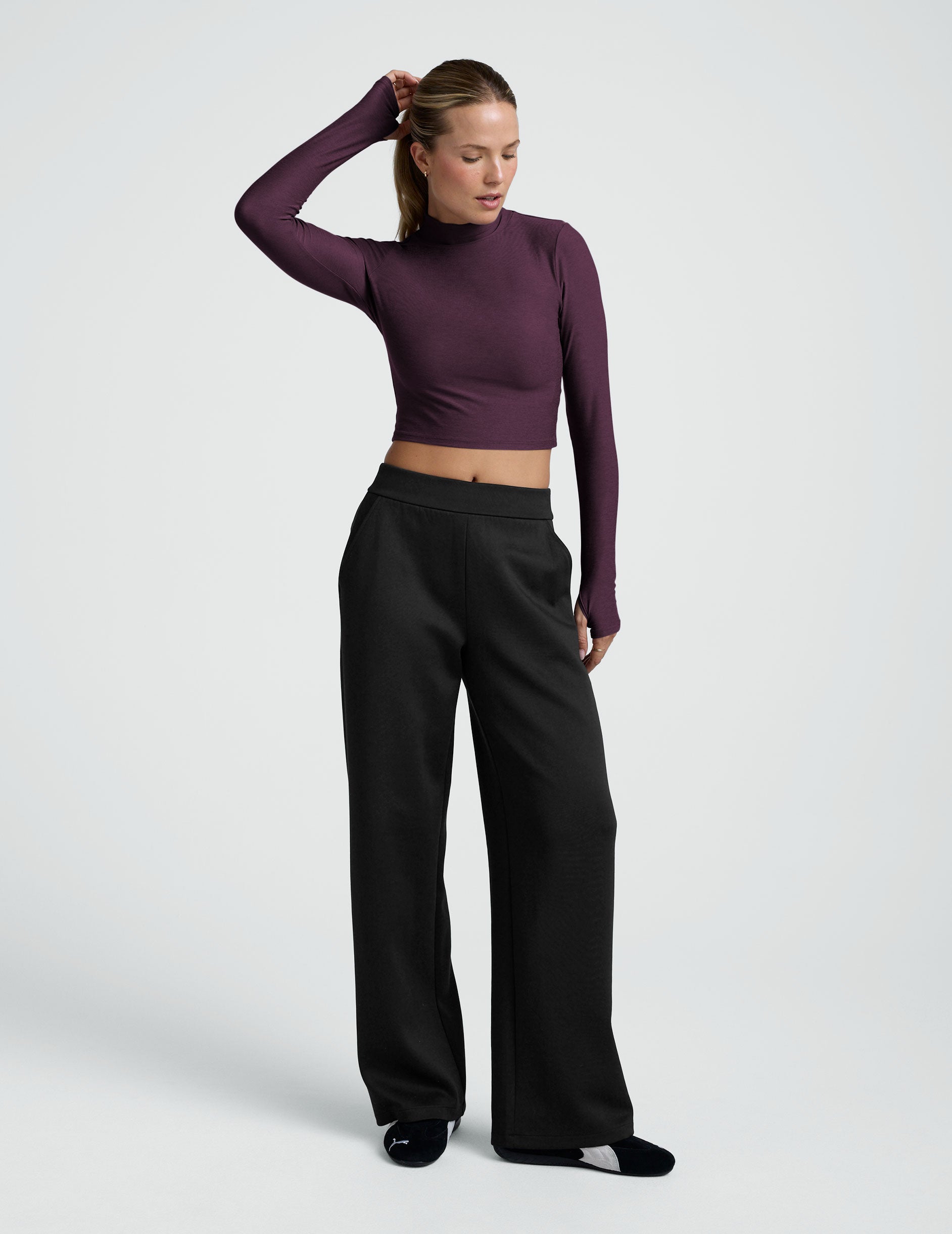 woman wearing a cropped purple long-sleeve mock-neck top and black pants on a plain background.