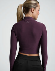 woman wearing a cropped purple long-sleeve mock-neck top and black pants on a plain background.