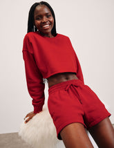 woman wearing a red pullover sweatshirt and sweat shorts against a plain background.
