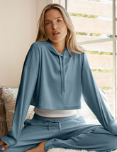 woman wearing a blue ribbed cropped hoodie and pants sitting in a room with a window in the background.