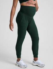 model wearing green maternity midi length joggers with pockets on a plain background.