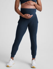 pregnant woman wearing navy blue maternity joggers with pockets on a plain background.