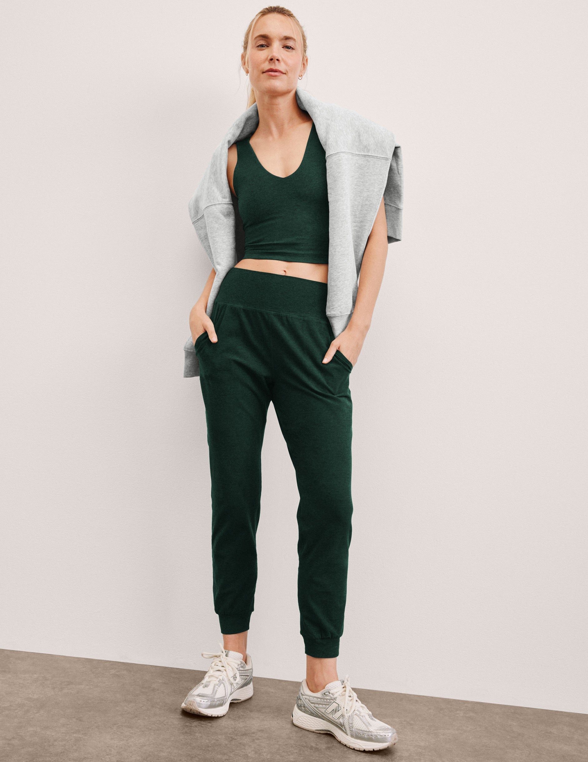 woman wearing a green cropped tank and joggers with a gray jacket against a plain background.