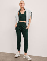 woman wearing a green cropped tank and joggers with a gray jacket against a plain background.