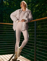 person wearing a nude quilted jacket and light pink leggings standing on a wooden deck with greenery in the background.