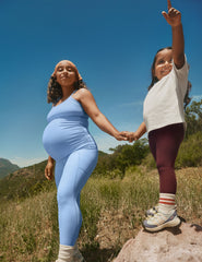 model wearing a blue maternity racerback tank and leggings and young girl in burgundy toddler leggings holding hands on a mountain trail.
