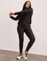woman wearing a brown long sleeve and high-waisted midi length pocket leggings with a plain background.