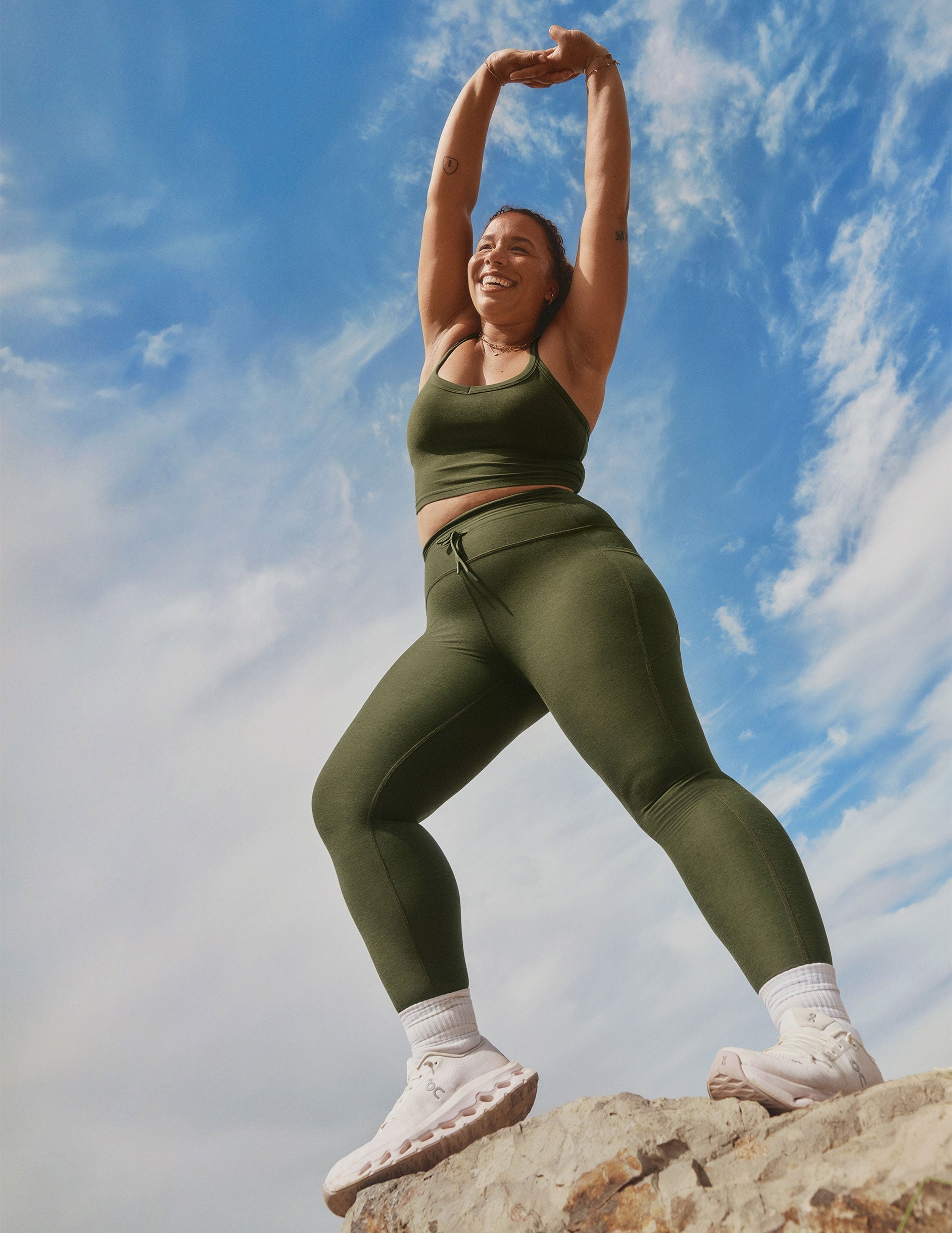 model in green cropped tank and leggings with a drawcord at waistband stretching outdoors against a blue sky.
