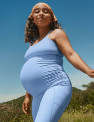 model in a blue maternity tank and leggings standing outdoors with a clear blue sky and greenery in the background.