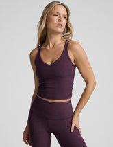 model is wearing a purple v-neck tank top with criss-cross strap back detail and purple high-waisted midi length leggings on a plain background.