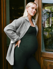 woman in a gray coat and green midi length jumpsuit standing in front of a glass door.