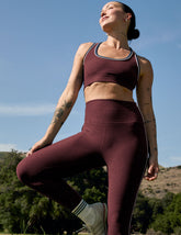 model in maroon sports bra with white contrast piping and leggings standing outdoors with a clear blue sky and greenery in the background.