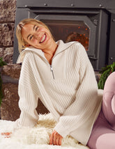 woman wearing a white quarter zip sweater sitting in front of a fireplace.
