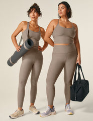 model is wearing a brown cropped racerback tank top and brown high-waisted midi leggings with a front crossover detail.