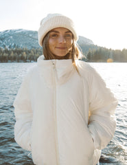 model wearing a white zip-up puffer jacket and blue leggings standing in a snowy landscape with mountains in the background.