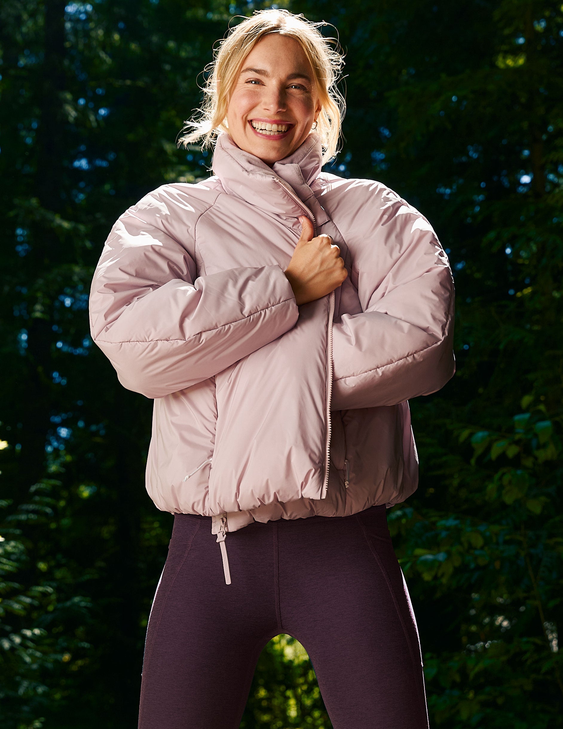 Person wearing a light pink puffer jacket and dark purple leggings standing against a green leafy background.