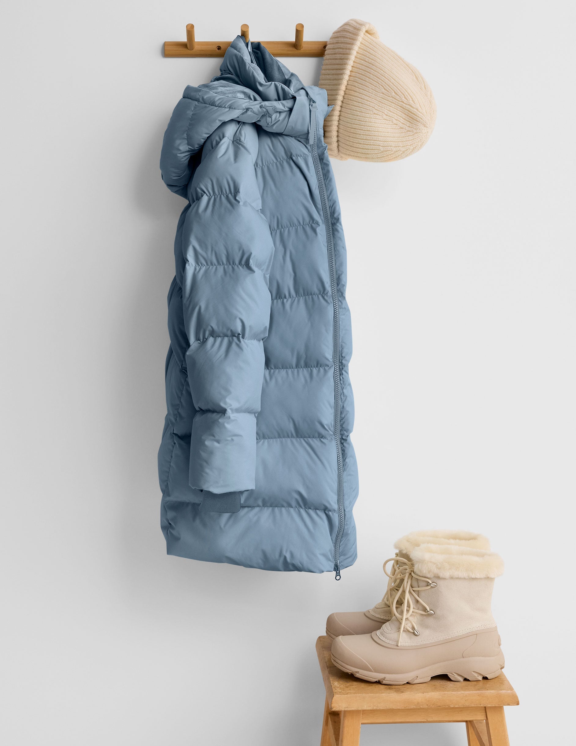 Blue puffer jacket, beige hat, and boots on a white wall with wooden hooks and stool.