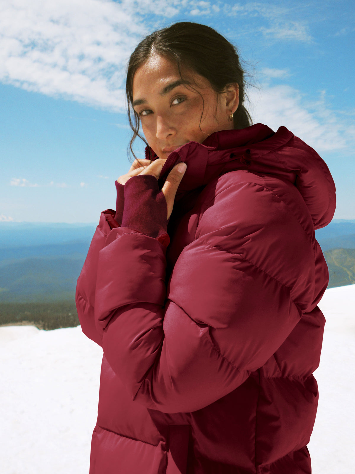 woman wearing a red mid thigh length puffer jacket with a hood against a snowy background.