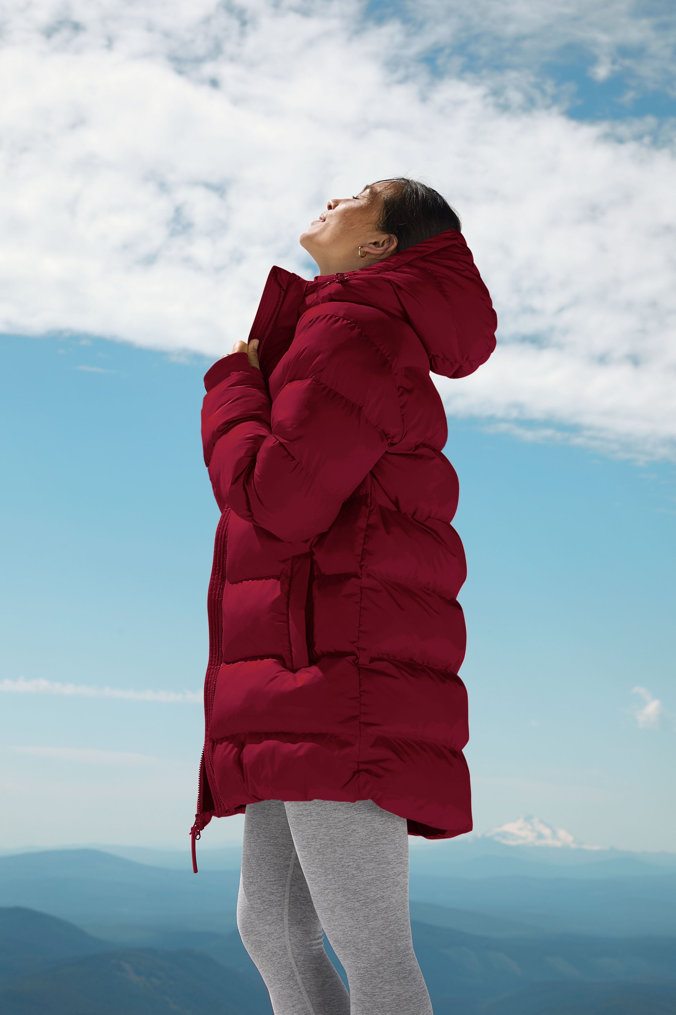 woman wearing a red mid thigh length puffer jacket with a hood against a snowy background.