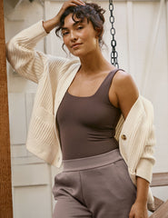 woman wearing a beige cardigan over a brown top and pants, standing in a neutral setting.