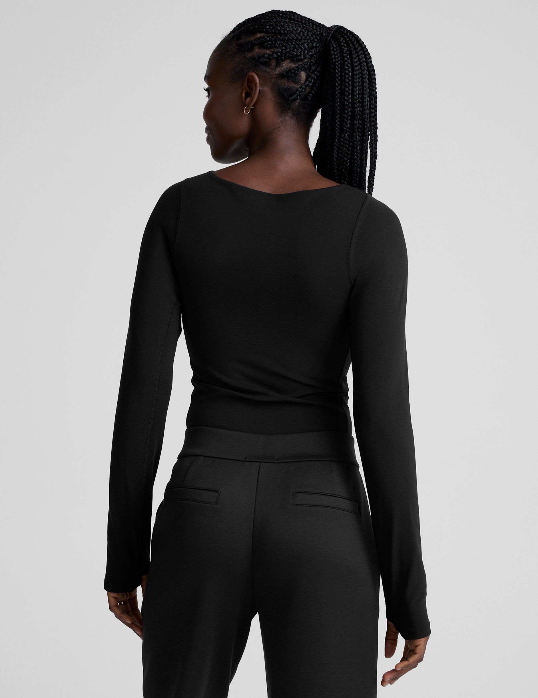 woman wearing a black long-sleeve boatneck bodysuit and black pants on a plain background.