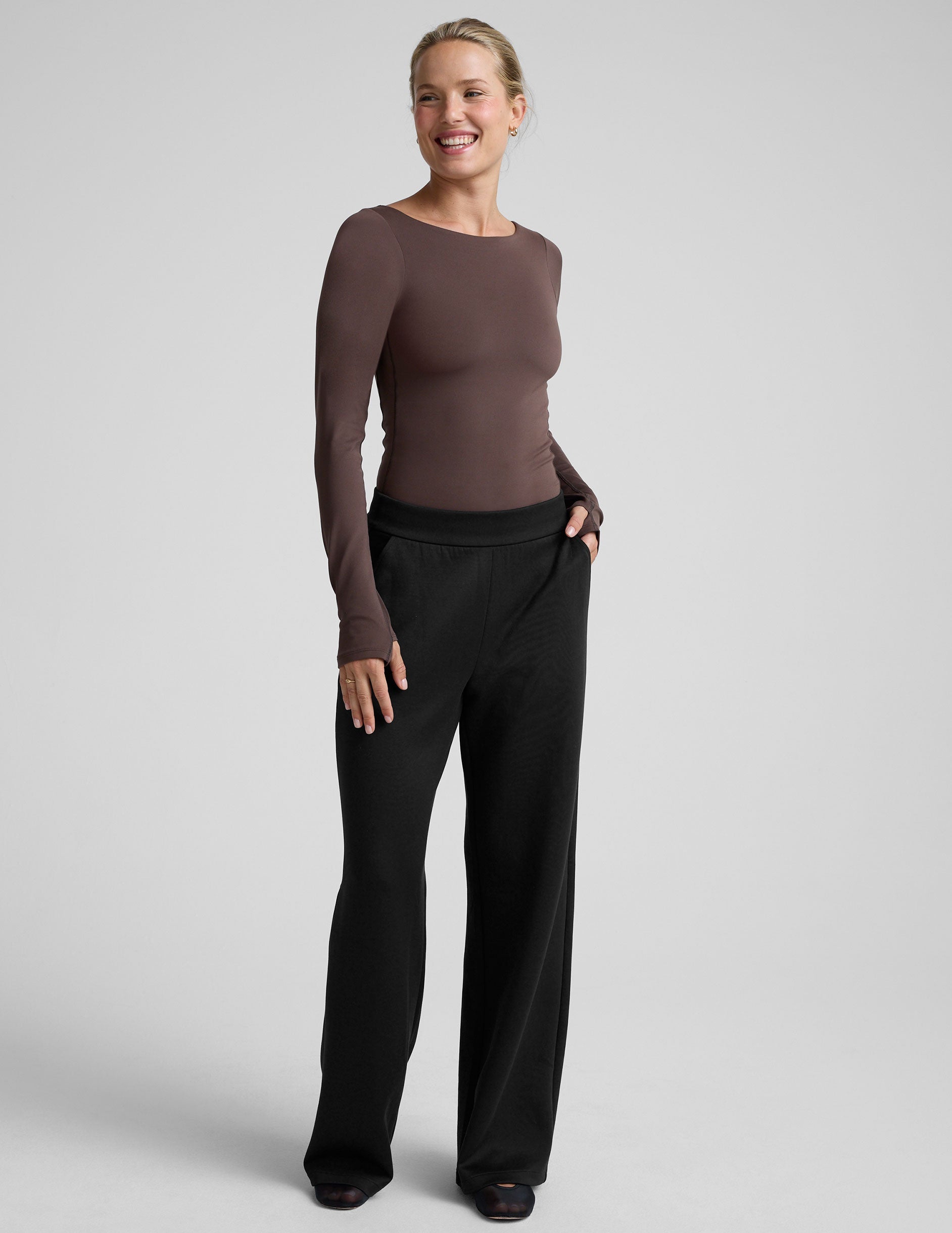 woman wearing a brown long-sleeve boat neck bodysuit and black pants against a plain background.