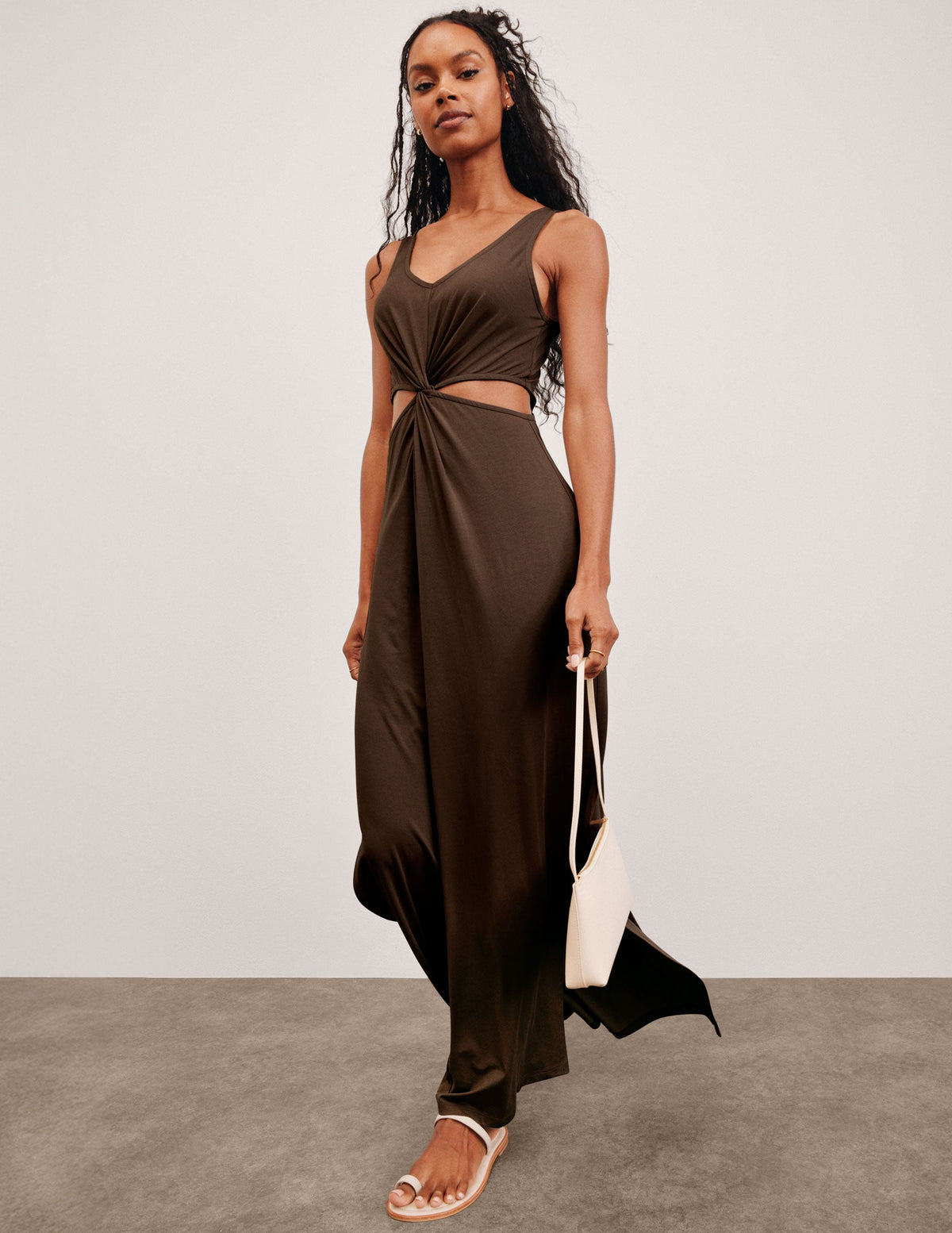 model wearing a brown dress with cut-out design, holding a white bag on a plain background. 