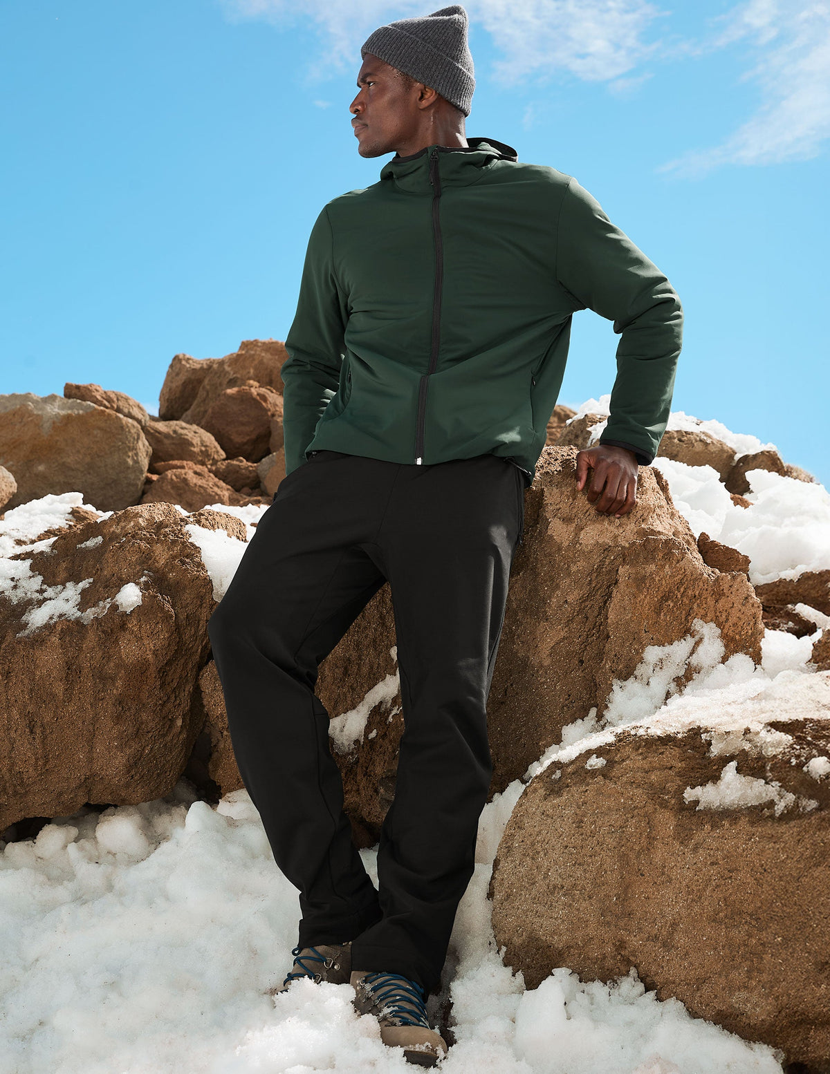 man wearing a green jacket, gray beanie, and black pants standing on snow-covered rocks with a blue sky.