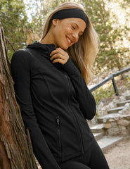 woman in black zip-up hooded jacket leaning against a tree outdoors.