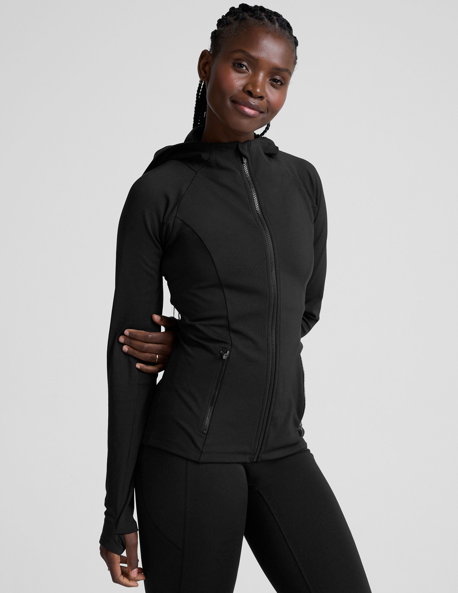 woman in black zip-up hooded jacket and leggings against a plain background.