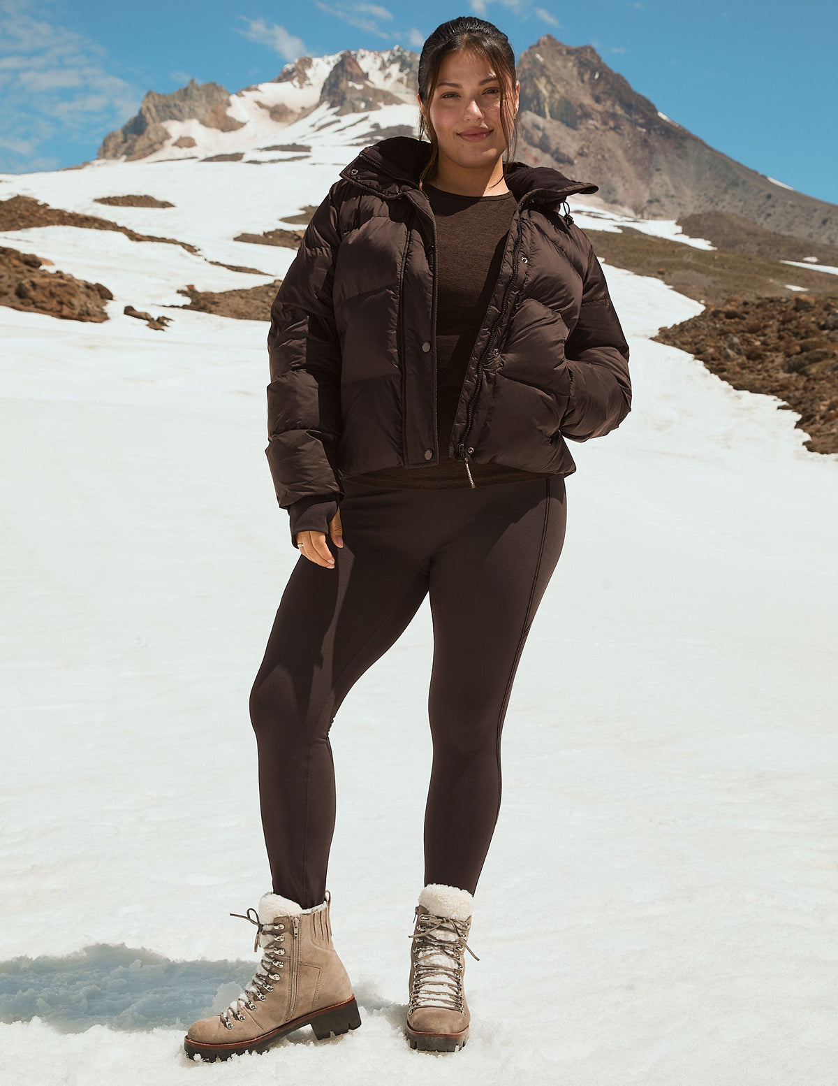 woman wearing a brown puffer jacket, brown leggings, and beige boots standing on a snowy mountain. 