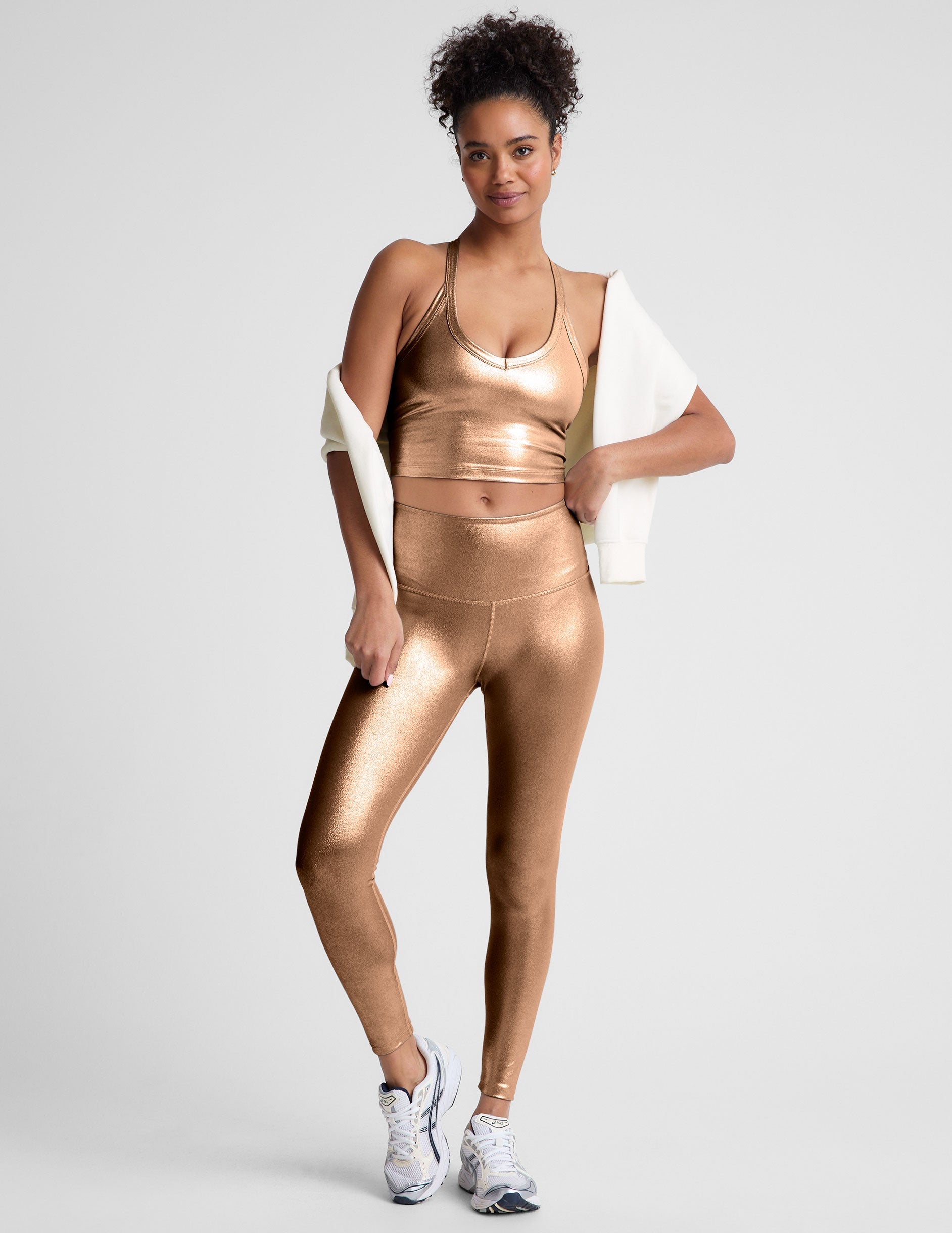 woman wearing a gold v-neck cropped tank and leggings on a plain background.