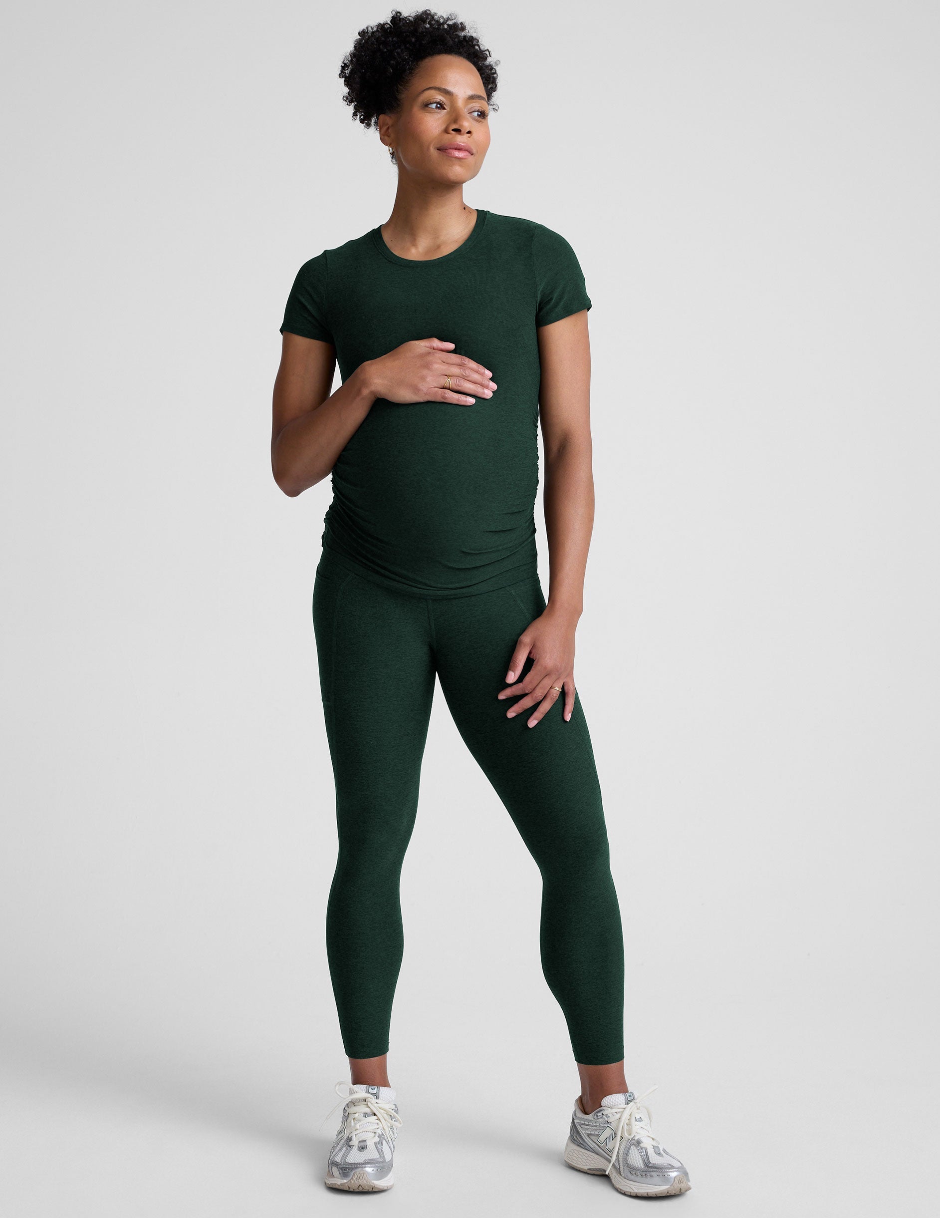 model woman wearing a dark green maternity t-shirt and leggings against a light gray background.