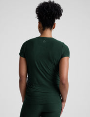 model woman wearing a dark green maternity t-shirt and leggings against a light gray background.