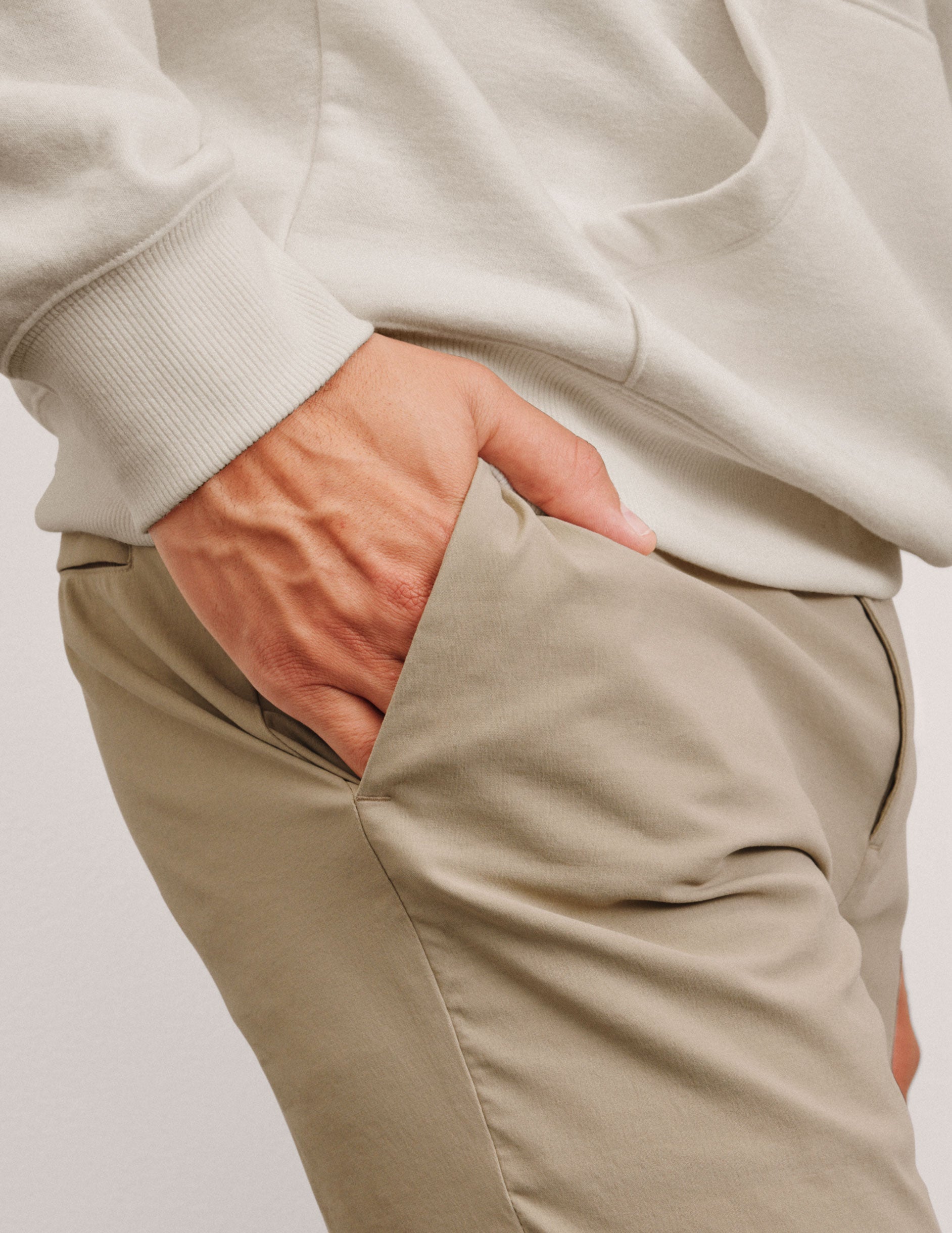 man wearing a beige sweater and pants with one hand in the pocket against a plain background.