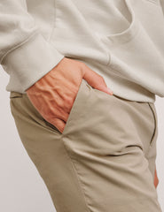 man wearing a beige sweater and pants with one hand in the pocket against a plain background.