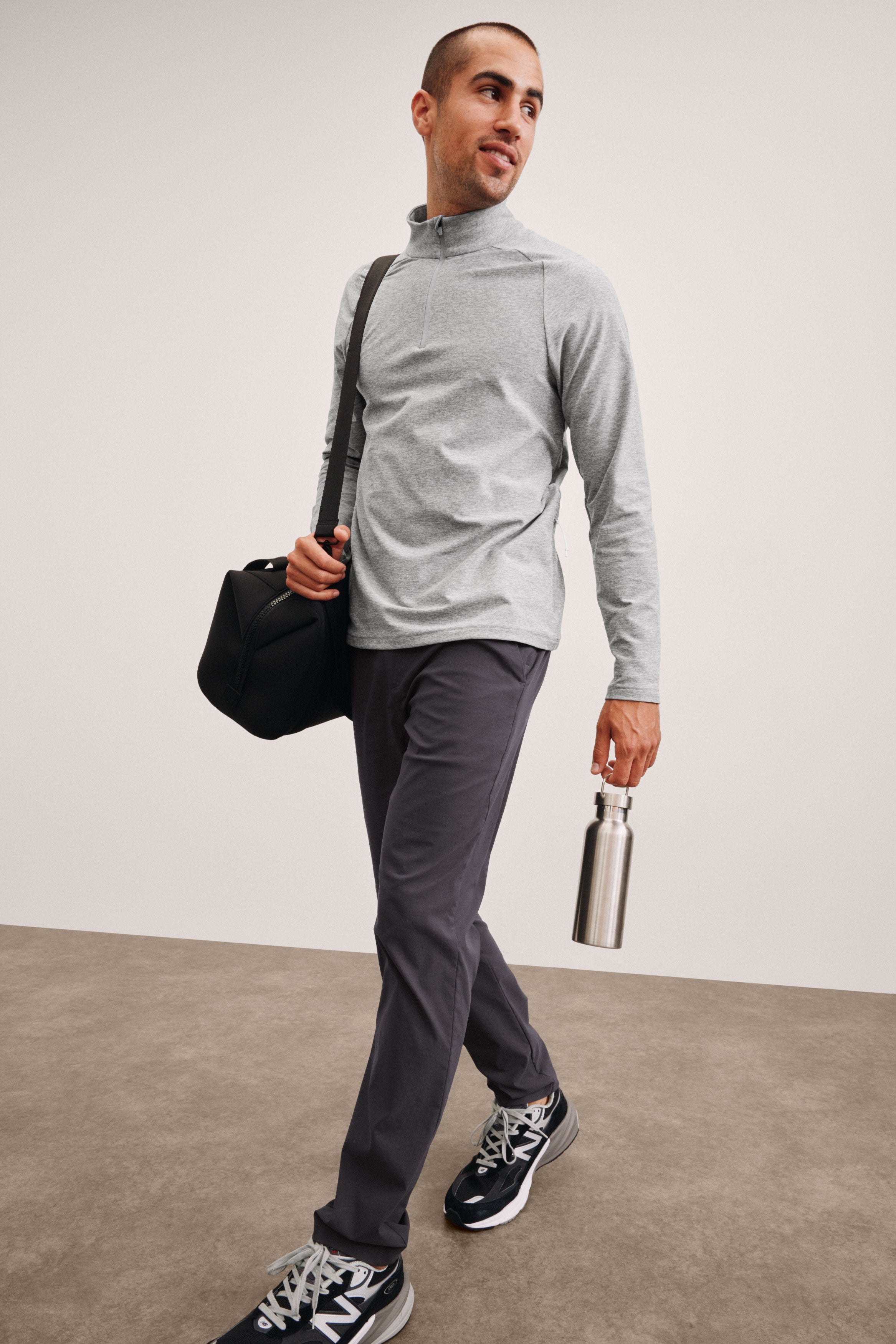 men's model in gray long-sleeve shirt and dark pants holding a black bag and silver water bottle against a plain background.