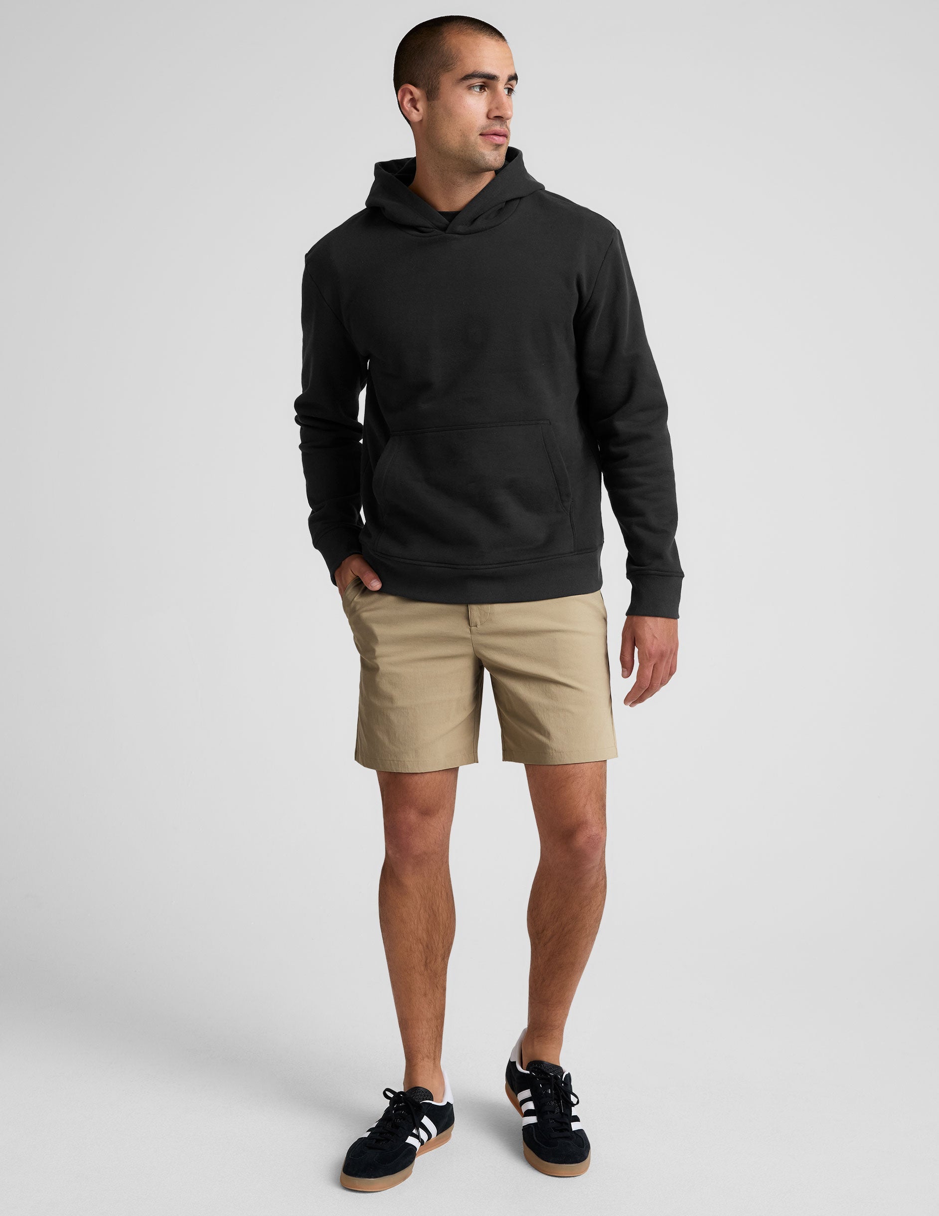 man wearing a black hoodie and beige chino shorts with pockets standing against a plain background.