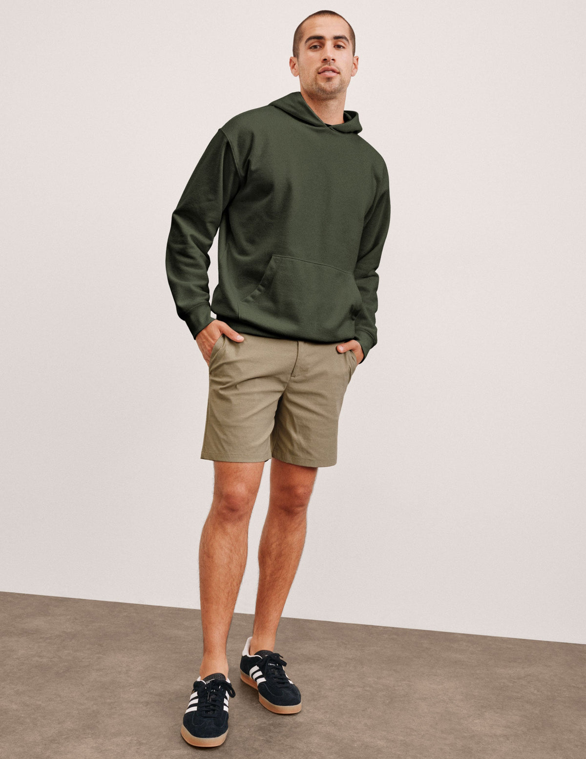 man wearing a green hoodie and beige chino shorts with pockets standing against a plain background.