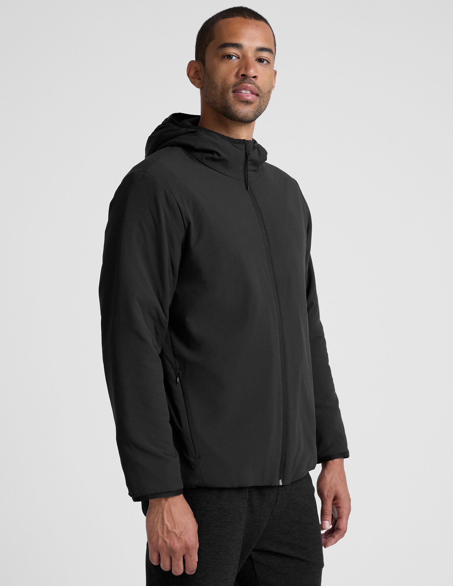 male model wearing a black zip-up jacket with a hood and pockets against a plain background.