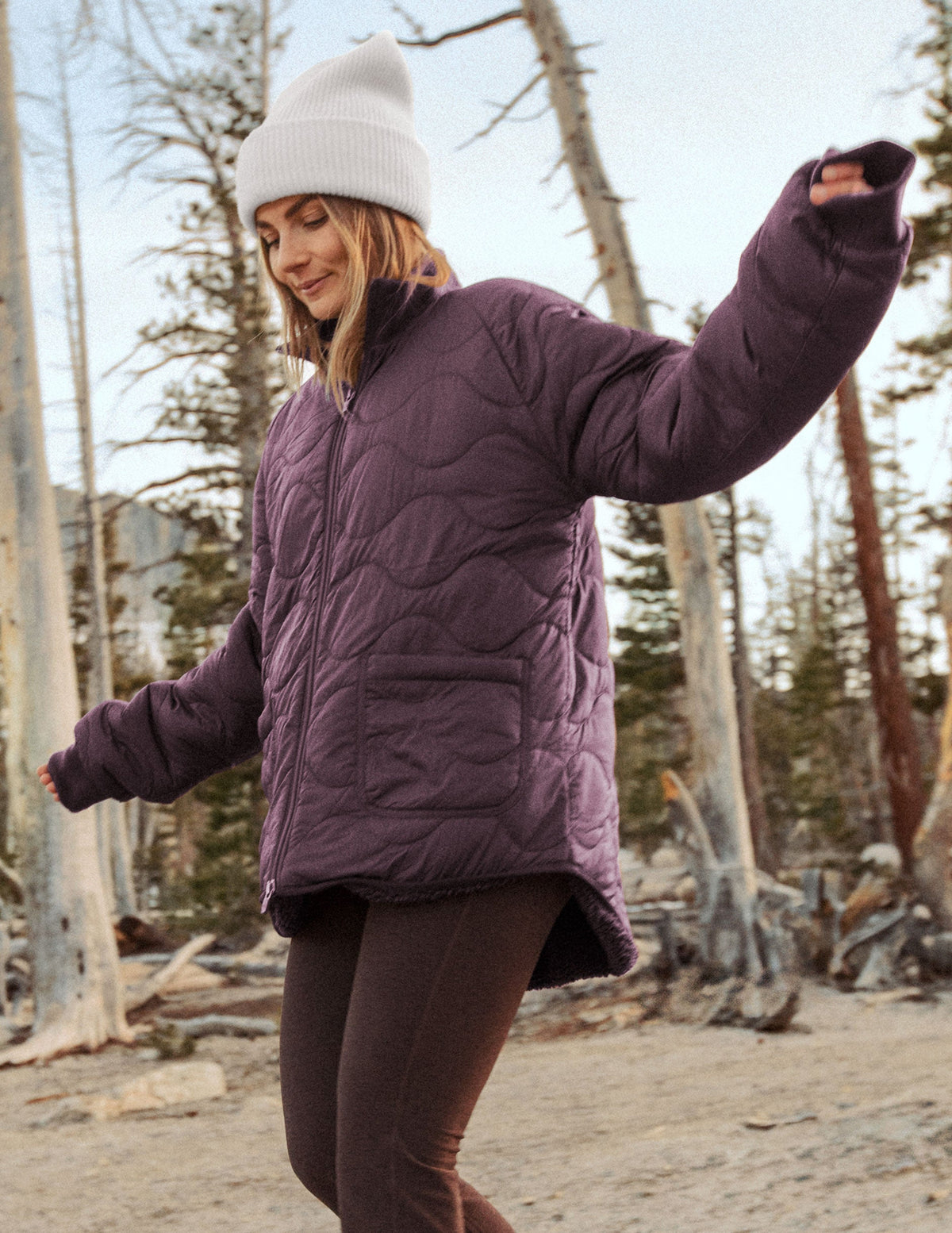 woman wearing a purple 2-way quilted jacket and white beanie in a forest setting.